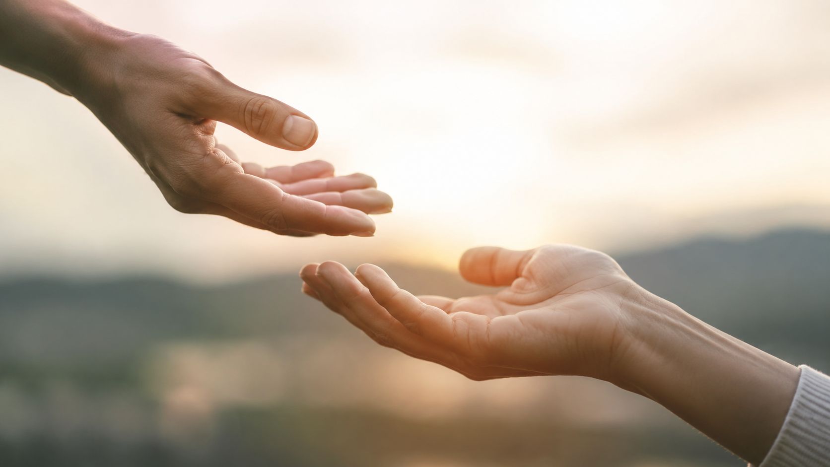 Two people reaching out with their hands towards each other against a soft, golden sunset background.